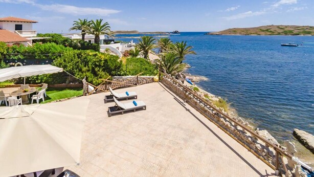 Un patio frente al mar con sillones de descanso, una mesa con sombrillas y vistas al océano y colinas distantes.