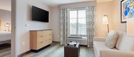 Une chambre d’hôtel comprenant un lit, un canapé, un téléviseur à écran plat, une armoire, une fenêtre avec des rideaux à motifs et une moquette colorée.