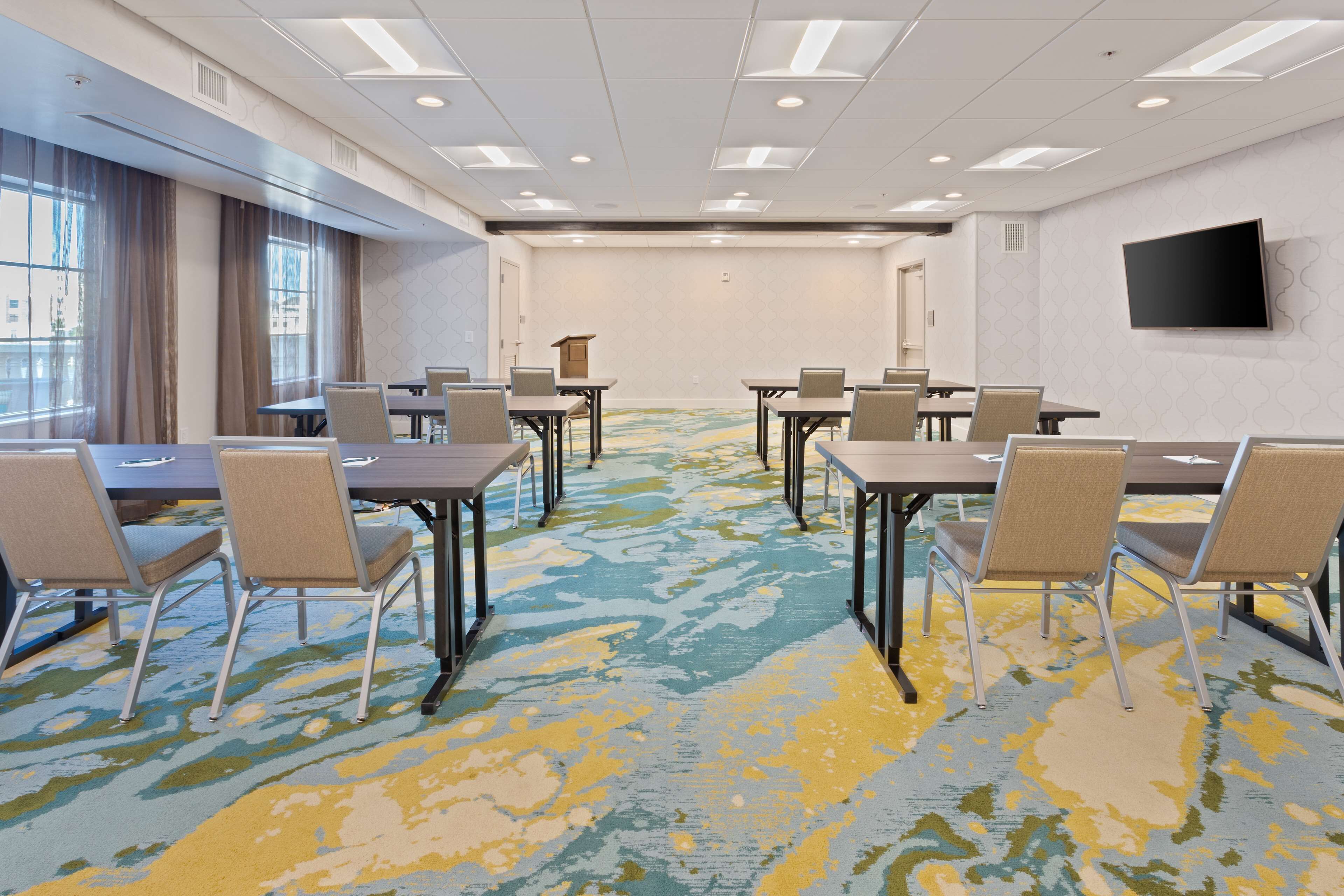 A conference room with a patterned carpet, tables, chairs, a flat-screen TV, and large windows with curtains.