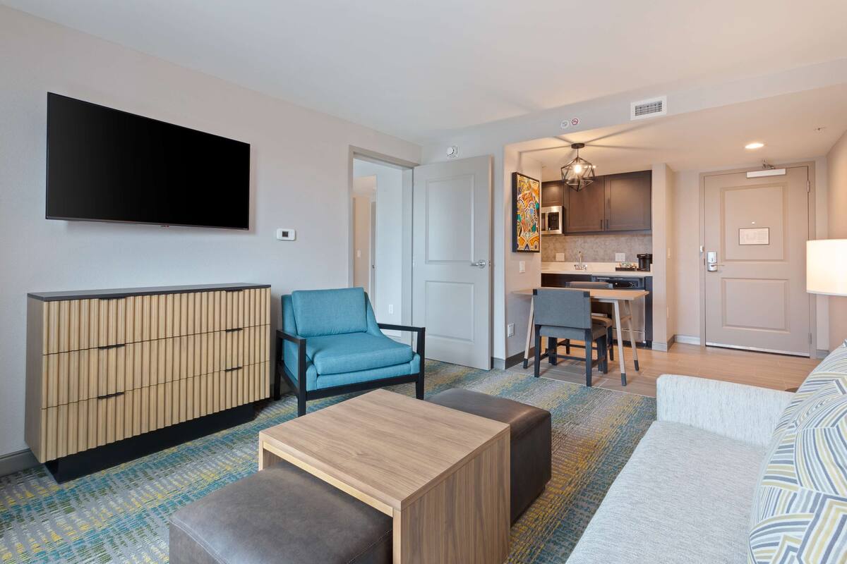 A modern living room with a flat-screen TV, a sofa, a blue armchair, a wooden coffee table, and a kitchen area with a dining table and chairs.