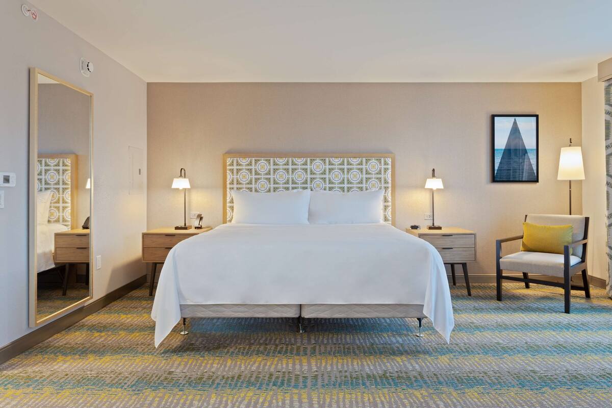 A hotel room with a large bed, two bedside tables, a chair, a mirror, and a framed picture on the wall.
