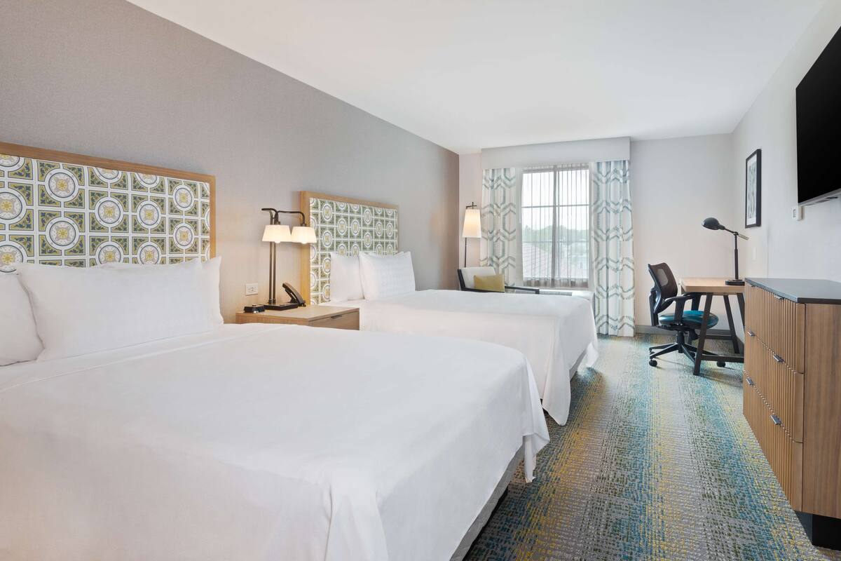 A hotel room with two beds, a desk, a chair, a TV, and a patterned headboard.