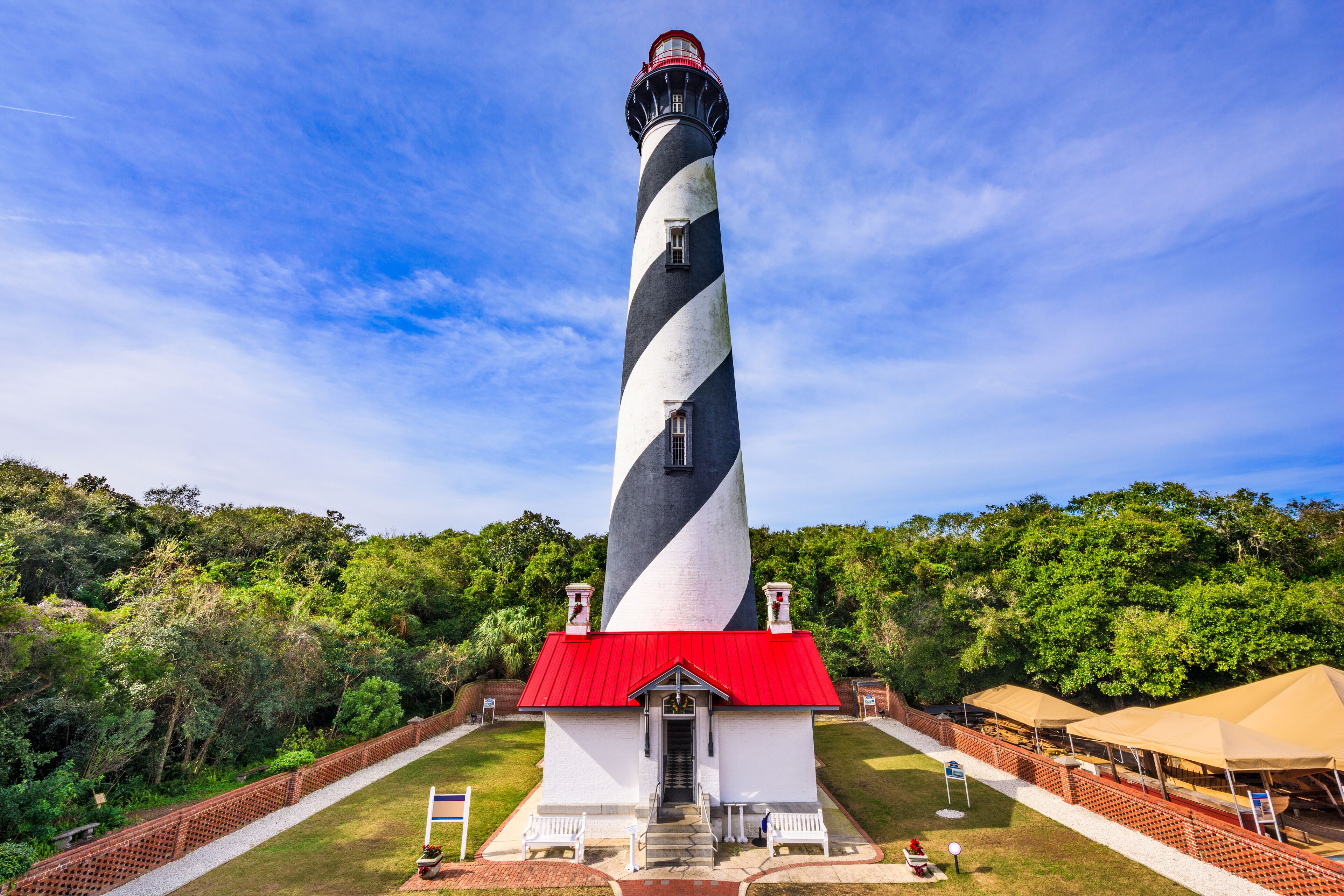 A lighthouse with a red roof and black and white stripes, surrounded by greenery and a clear sky.