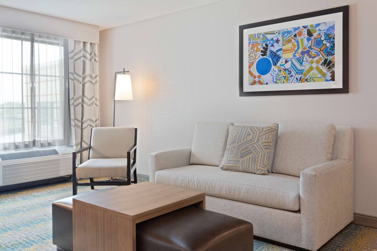 A modern hotel room with a beige sofa, a wooden coffee table, a patterned armchair, and a large abstract painting on the wall.