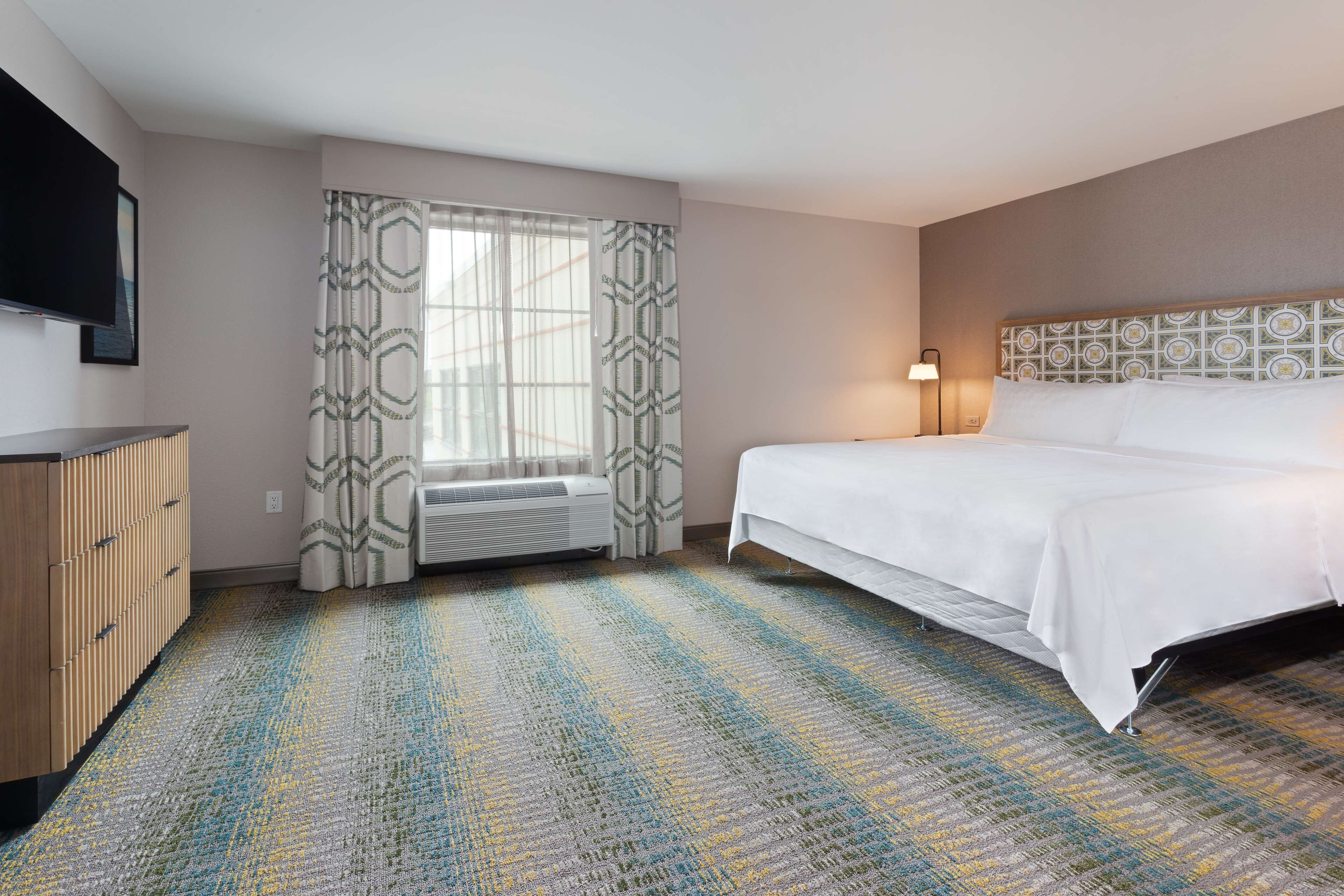 Habitación de hotel con cama, televisor de pantalla plana montado en la pared, ventana con cortinas estampadas, calefactor portátil y alfombra con diseño.