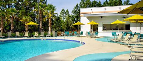 A swimming pool with lounge chairs and yellow umbrellas, surrounded by palm trees and a building with a flat roof.