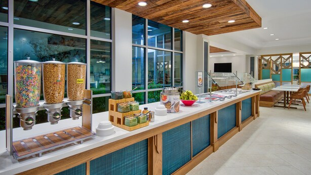 A breakfast bar with various food dispensers, a seating area with a table and chairs, and large windows with a view of greenery.