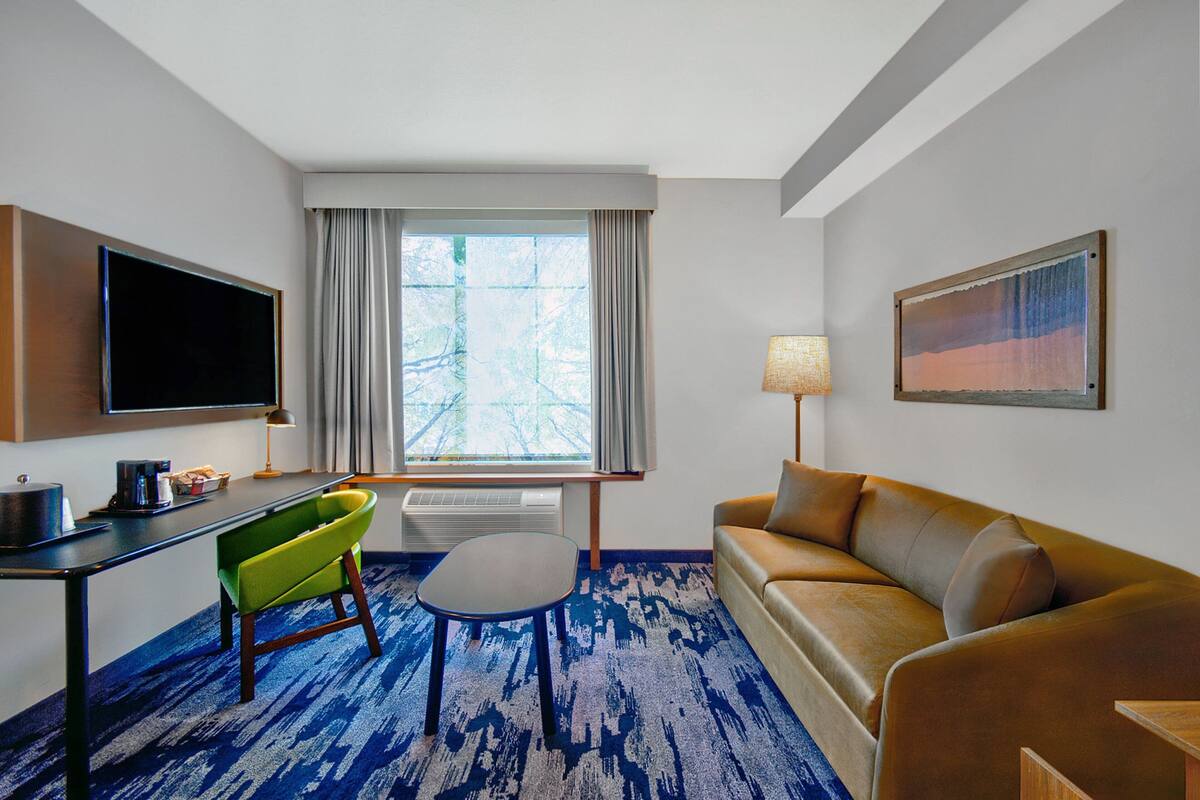 A hotel room with a flat-screen TV, a sofa, a chair, a small table, a lamp, and a painting on the wall.
