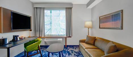 Quarto de hotel com televisão de tela plana, sofá, poltrona, mesa pequena, abajur e um quadro na parede.