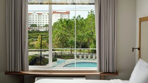 A hotel room with a large window overlooking a swimming pool and a landscaped area with trees and a building in the background.