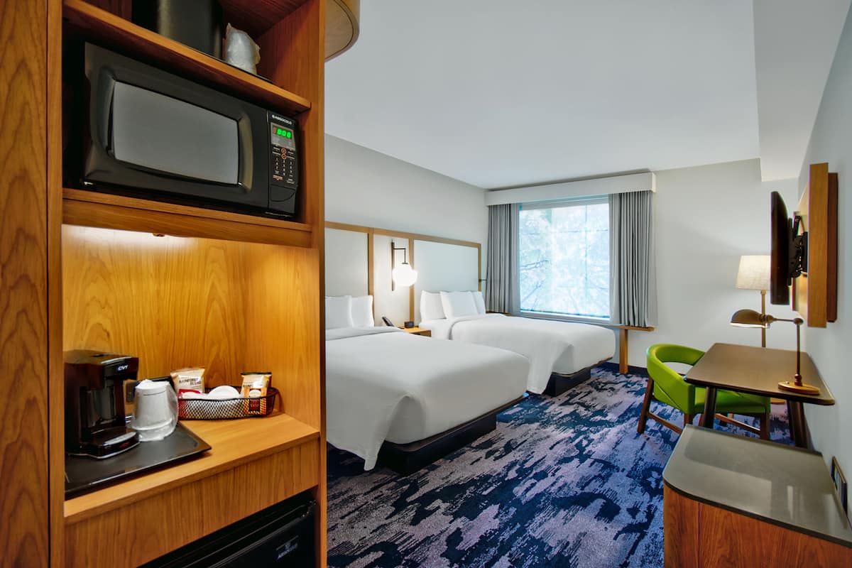 A hotel room with two beds, a microwave, a coffee maker, a desk with a lamp, and a view of the outdoors.