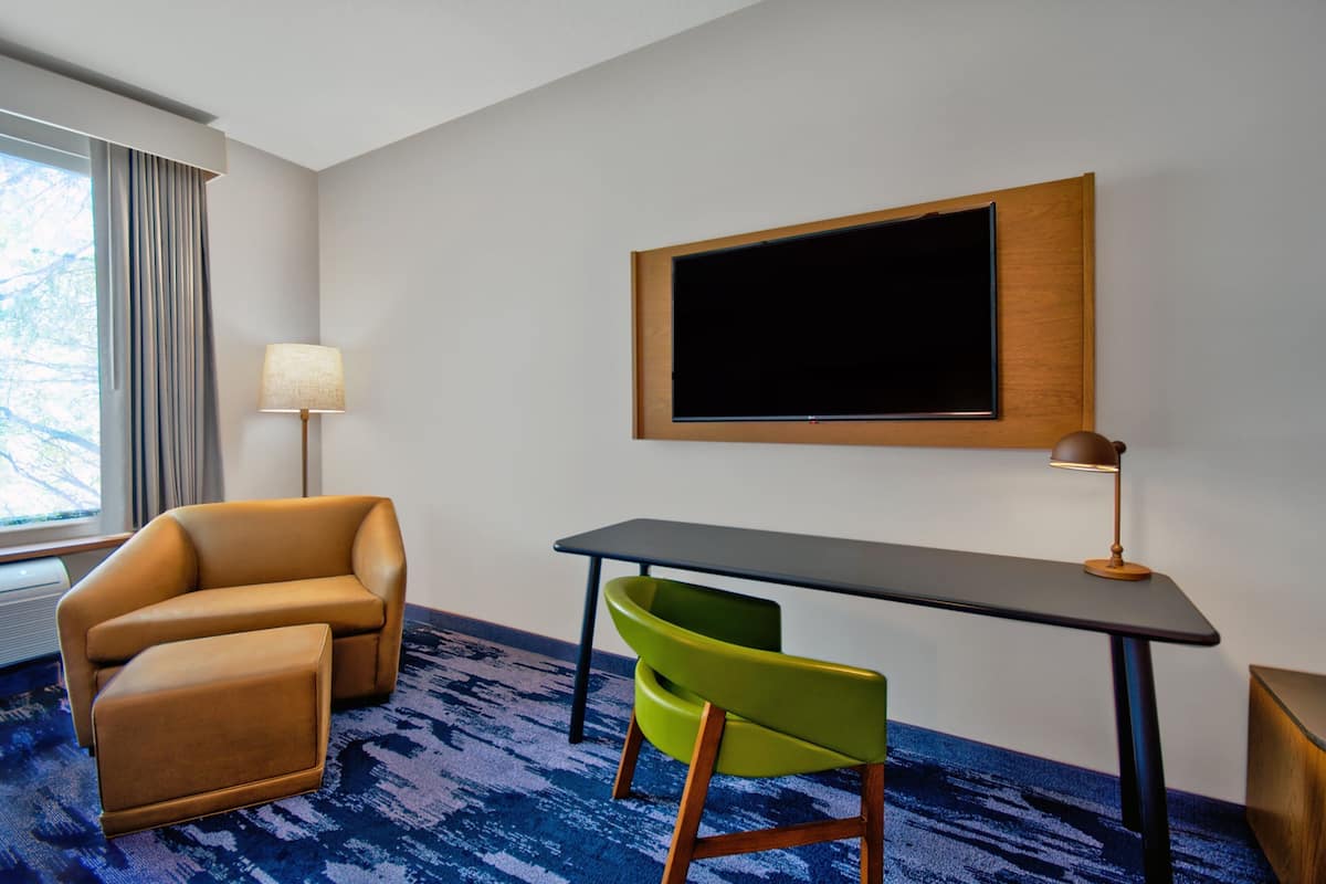 A hotel room with a sofa, a small table, a green chair, a lamp, and a TV mounted on the wall.