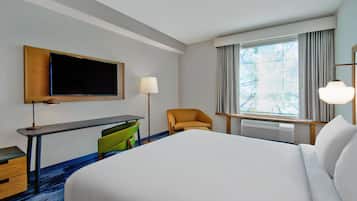 Quarto de hotel com uma cama grande, uma mesa com cadeira, televisão, luminária e janela com cortinas.