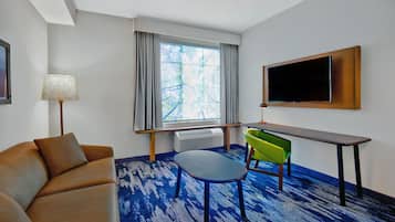 Quarto de hotel com sofá, mesa pequena, cadeira, televisão e janela com cortinas.