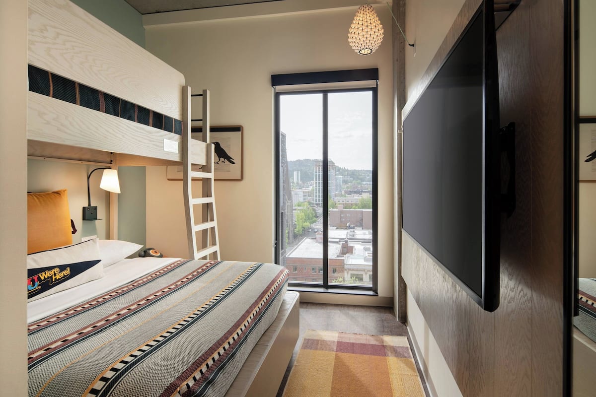 A bedroom with a bunk bed, a flat-screen TV, a window with a city view, a ladder, and a patterned rug.