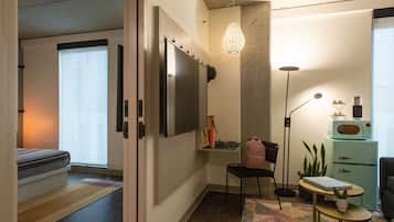 A compact living space with a bed, a desk with a computer, a chair, a small table, a lamp, and a refrigerator.