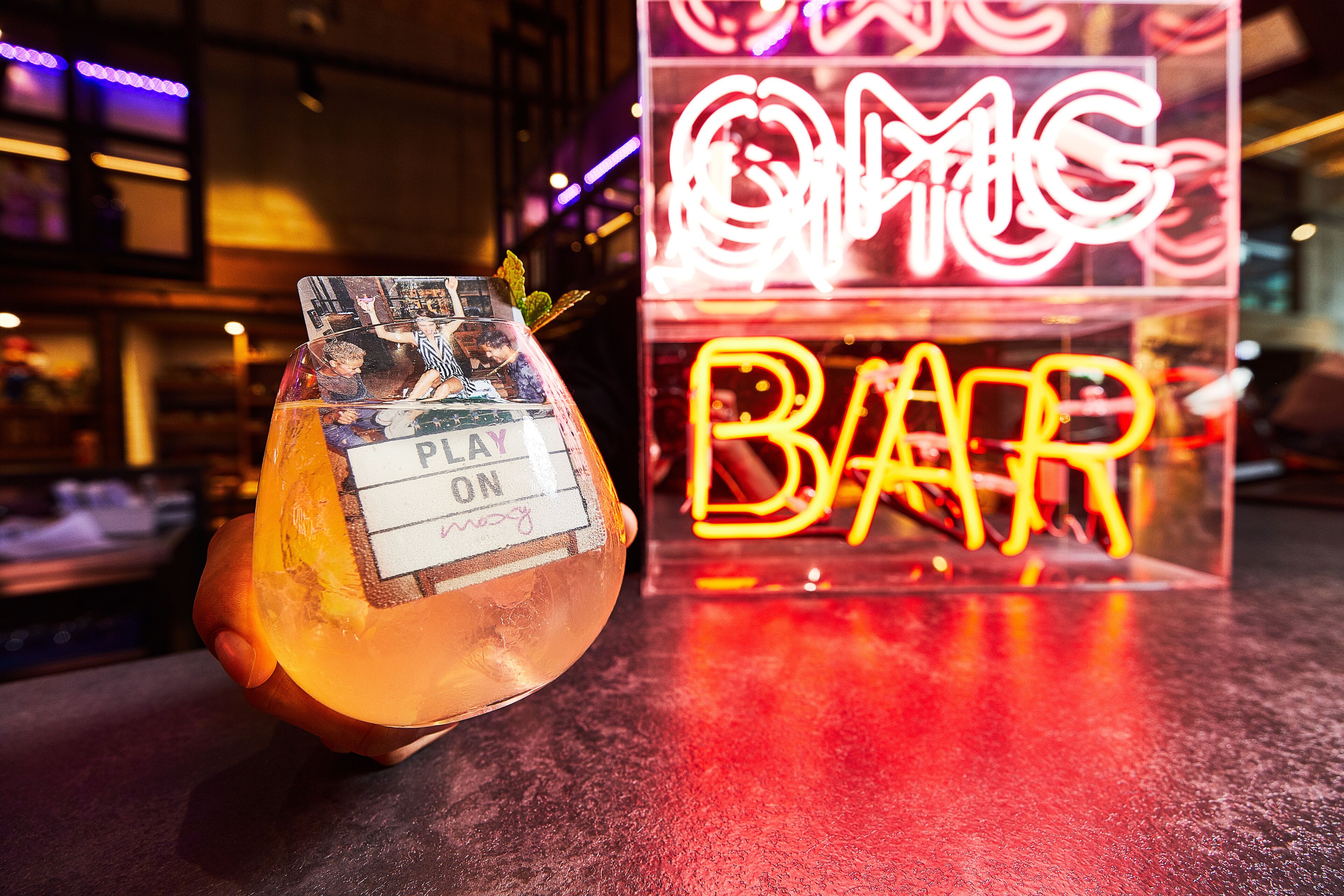 A cocktail in a glass with a label that reads "PLAY ON" and a neon sign in the background that says "OMG BAR".
