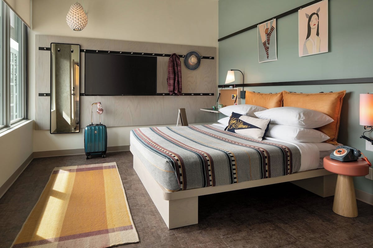 A modern hotel room with a large bed, a bedside table with a lamp, a suitcase, and artwork on the wall.