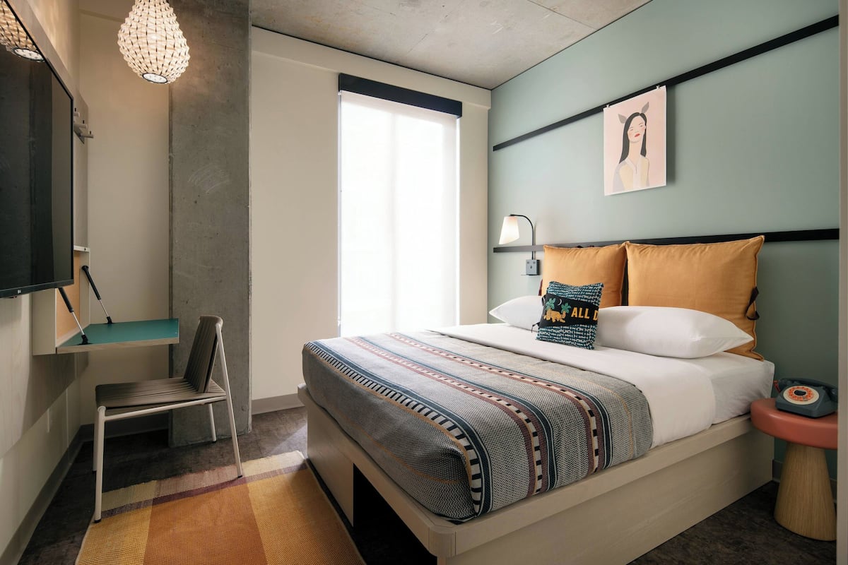A modern bedroom with a bed, a desk, a chair, a lamp, and a wall-mounted artwork.