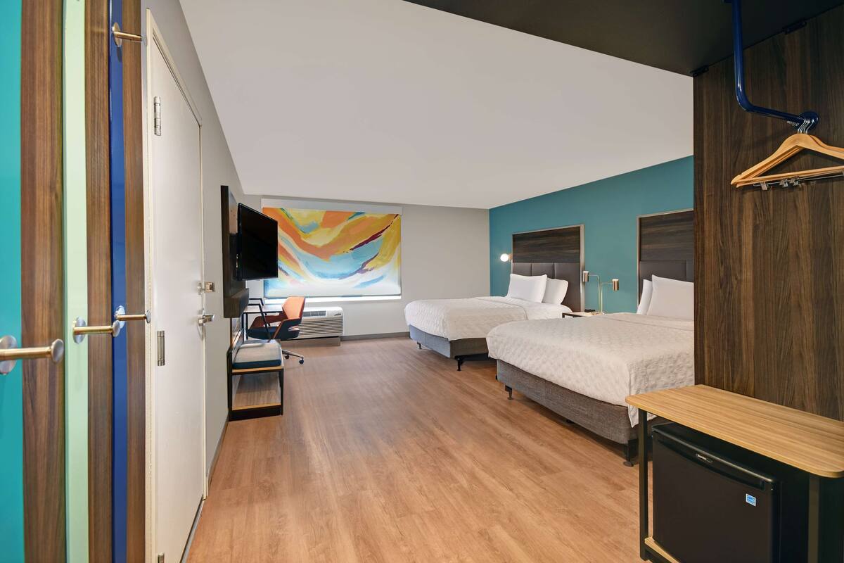 A hotel room with two beds, a desk, a TV, and a colorful abstract painting on the wall.