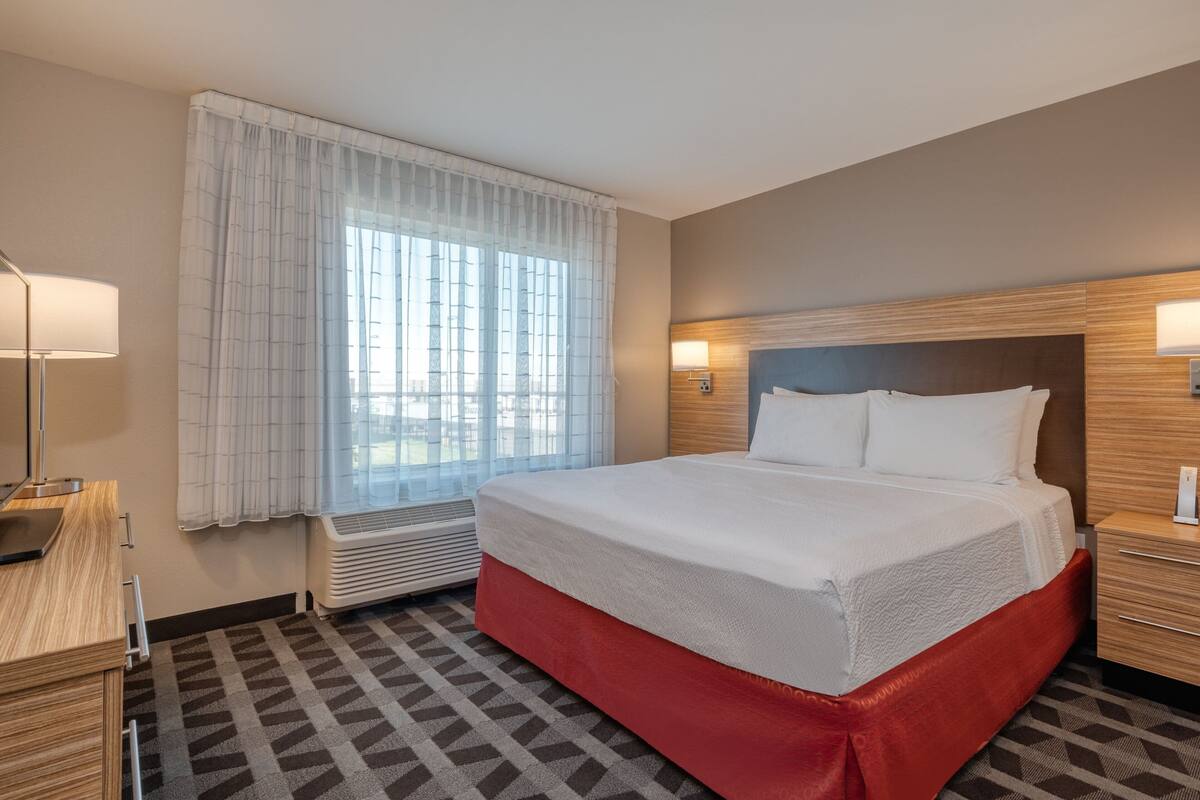Una habitación de hotel con una cama grande, una mesita de noche, una lámpara, una ventana con cortinas y un calefactor de pared.