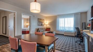 Et hotelværelse med spisebord, stole, sofa, fjernsyn og et vindue med gardiner.