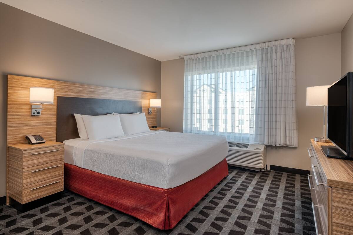 Habitación de hotel con una cama grande, una mesita de noche con un teléfono, un televisor y una ventana con cortinas.