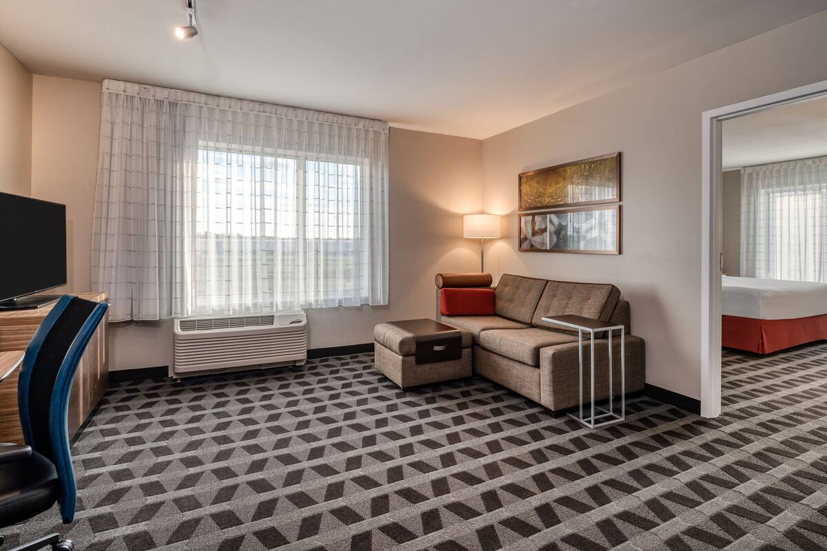 Habitación de hotel con sofá, escritorio, silla, lámpara y un amplio ventanal con cortinas.