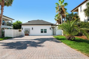 A residential house with a paved driveway, a white exterior, and a small front yard with palm trees.