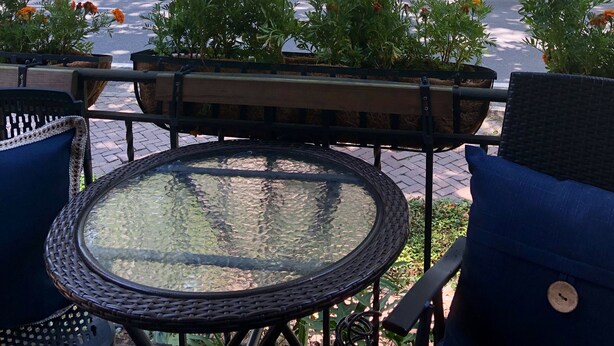 An outdoor seating area with wicker chairs and a glass-top table, surrounded by trees and a grassy park. - Forsyth Executive Rental (Savannah)