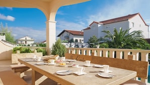 A sunlit outdoor dining area with a table set for a meal, surrounded by a white railing and overlooking a residential area with houses and greenery. - Luxury Villa Rosalia (Kastela)