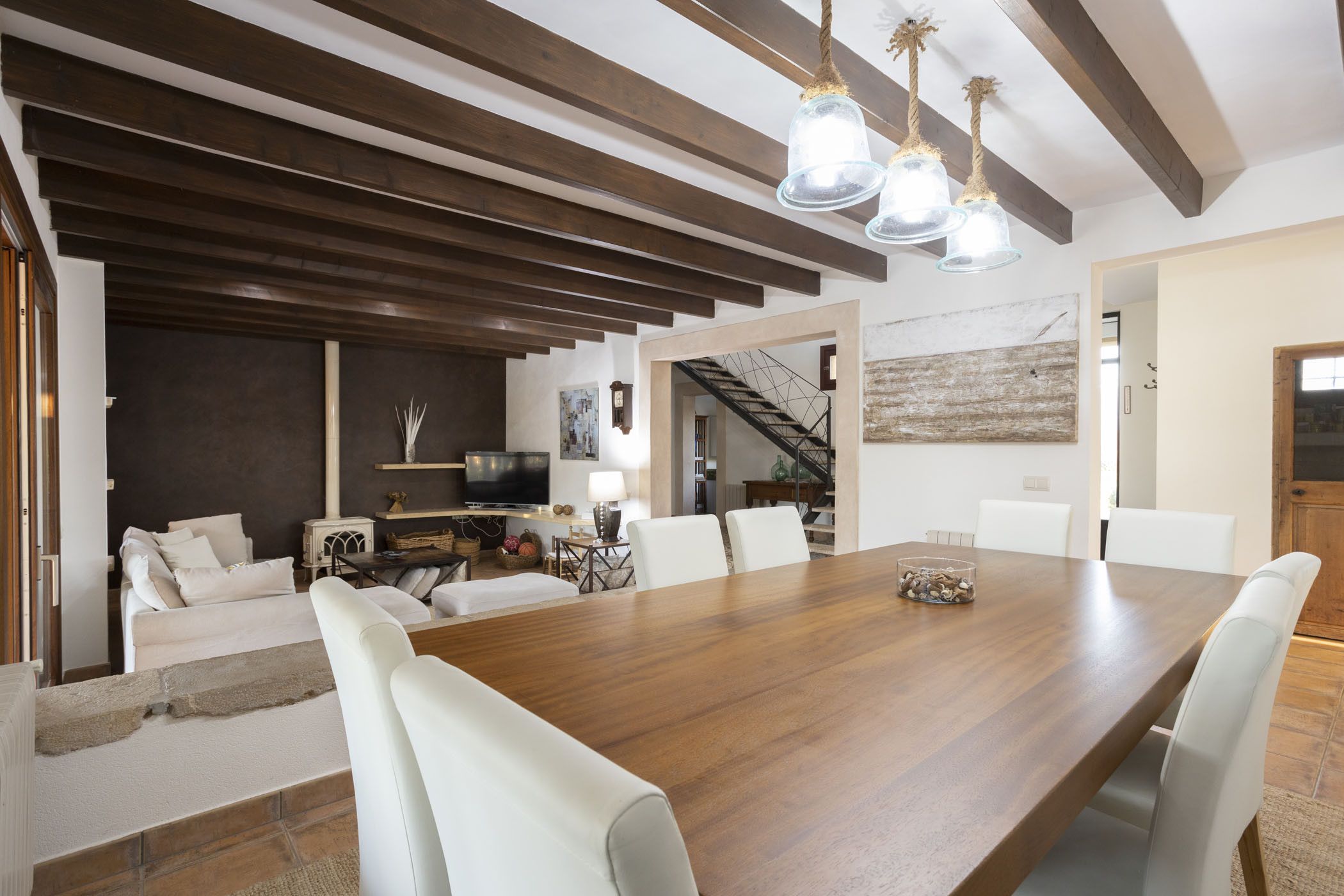 Un comedor amplio con una gran mesa de madera y sillas blancas, una zona de estar acogedora con un sofá, y una escalera que sube a un nivel superior.