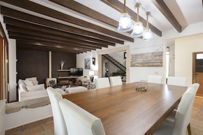 Un vaste espace à manger avec une grande table en bois et des chaises blanches, un coin salon confortable avec un canapé, et un escalier menant à un niveau supérieur.