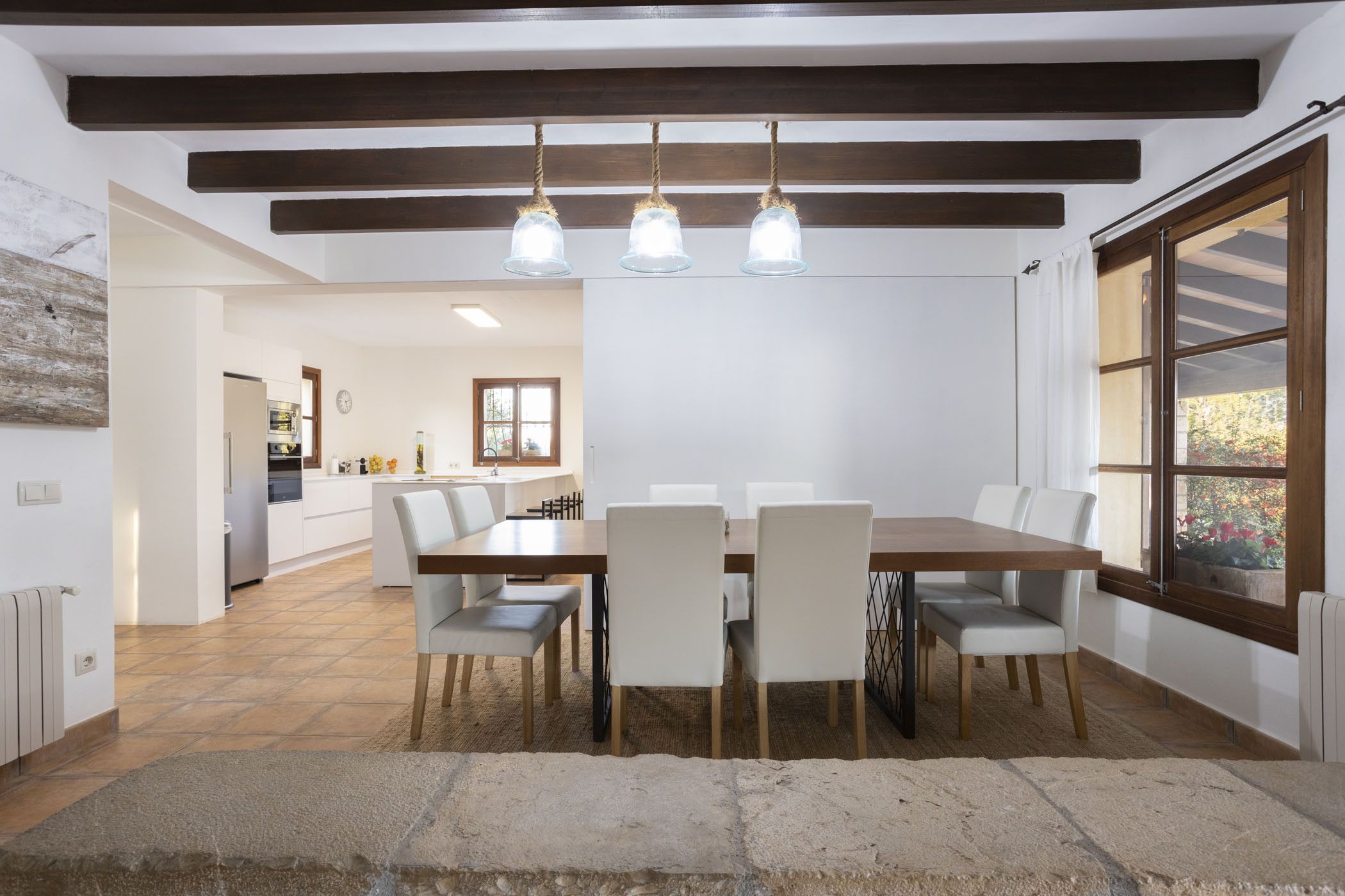Un comedor con mesa y sillas de madera, piso de piedra y vista a la cocina al fondo.