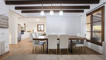 Un comedor con mesa y sillas de madera, piso de piedra y vista a la cocina al fondo.
