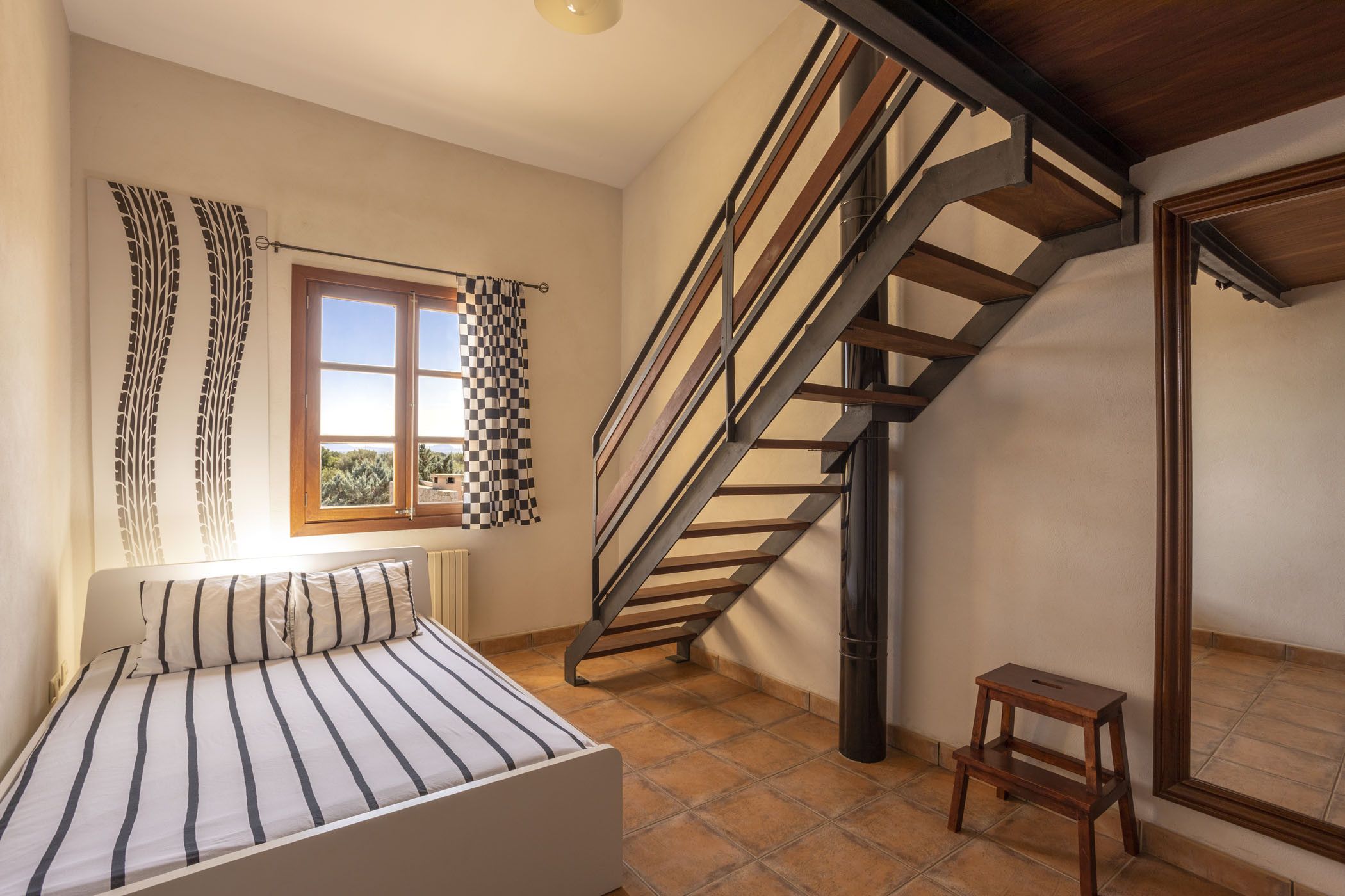 Un dormitorio con cama, una ventana con cortinas, una escalera de madera y un taburete pequeño de madera.