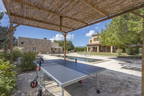 Une table de ping-pong installée à l’extérieur, sous une pergola en bois, entourée d’une piscine et de bâtiments en pierre.
