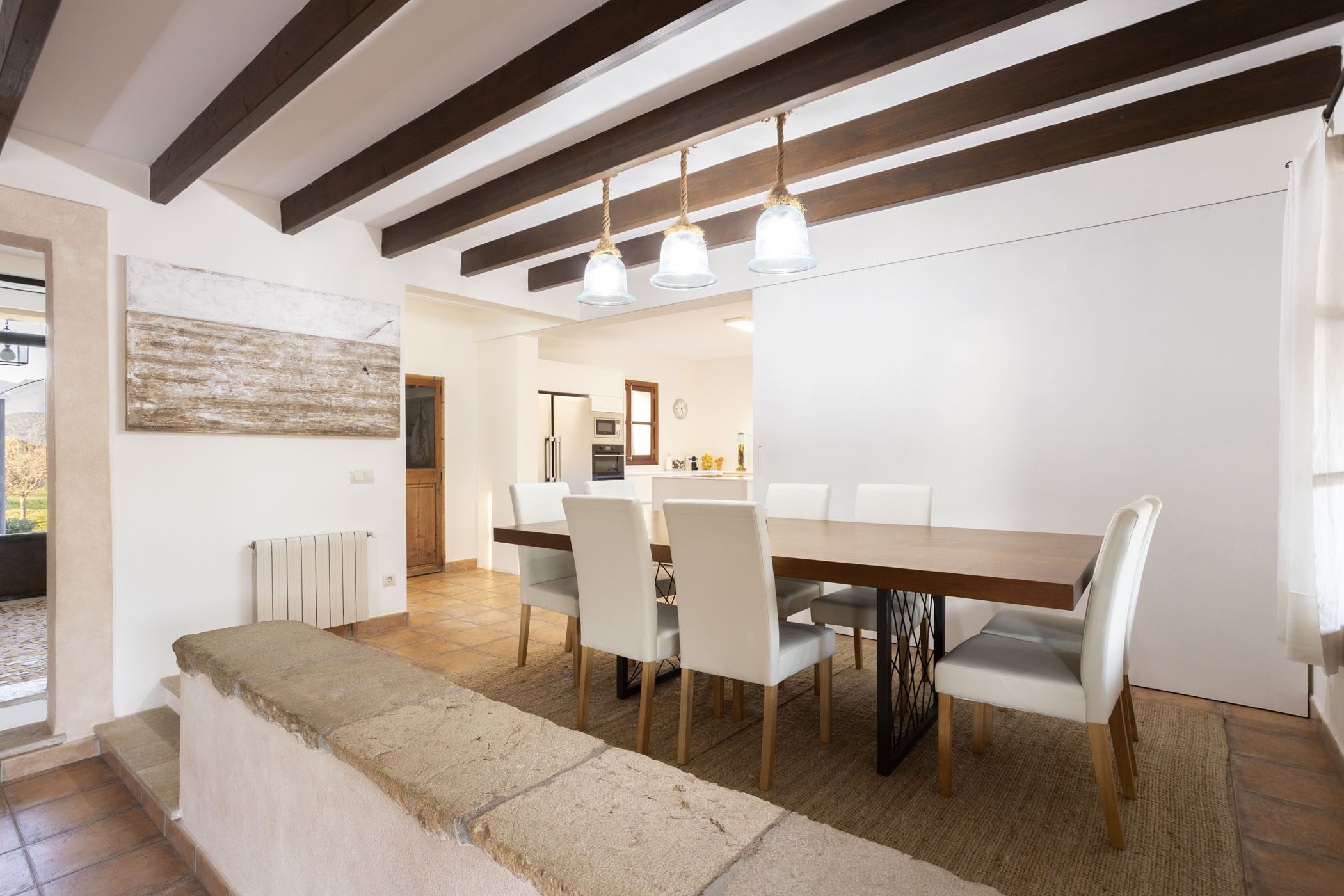Un comedor con mesa de madera, sillas blancas, un banco de piedra y una lámpara colgante.