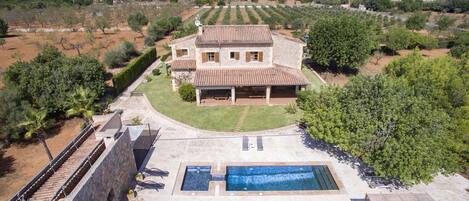 A large stone house with a swimming pool, surrounded by a well-maintained garden and olive groves.
