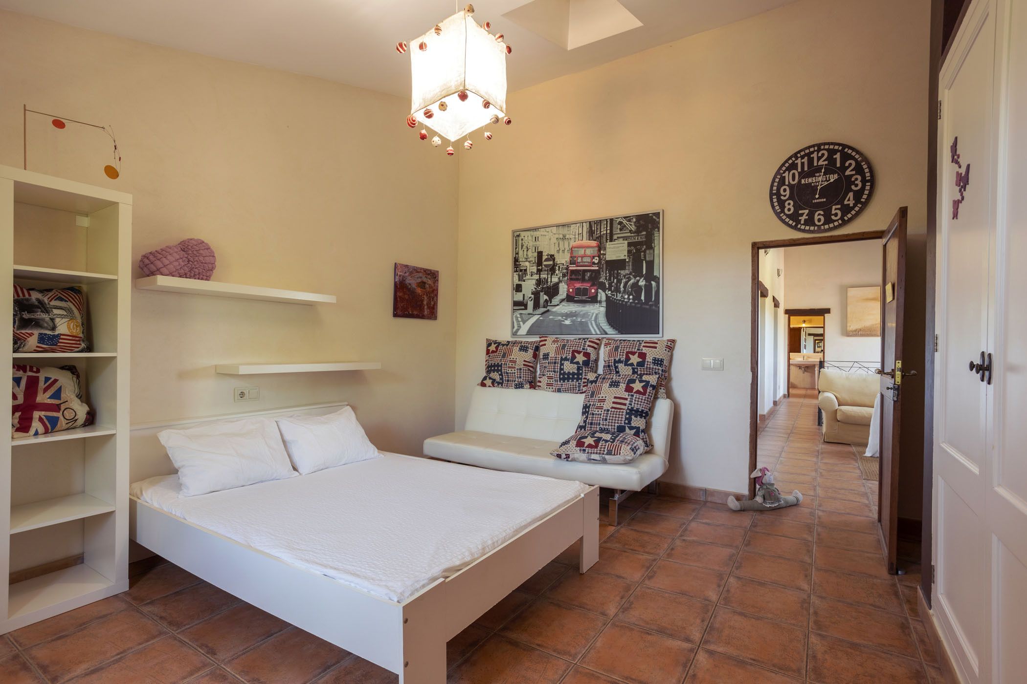 Un dormitorio con cama, un armario blanco, una estantería con almohadas, un reloj de pared y un pasillo que conduce a otra habitación.