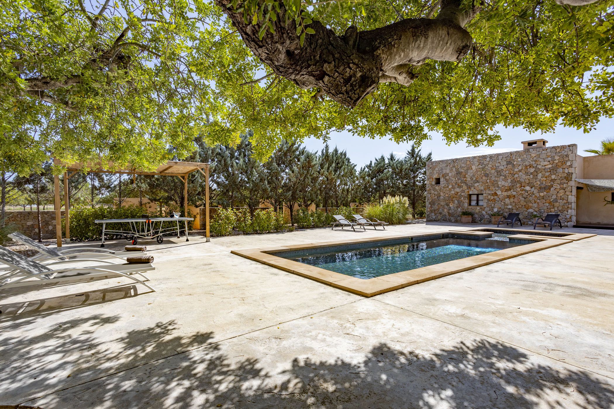 Una piscina con pared de piedra, sillas de descanso y una pérgola con mesa y sillas.