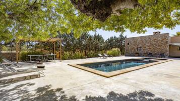 Una piscina con pared de piedra, sillas de descanso y una pérgola con mesa y sillas.