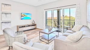 A modern living room with a sofa, armchairs, a coffee table, and a TV. There is a balcony with a view of trees and a railing. - Upstay - Condo w Pool and More - Near Disney (Winter Garden)
