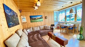 A wooden-paneled room with a large L-shaped sectional sofa, a wooden coffee table, and a dining area with a table and chairs.