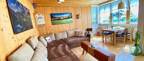 Una habitación con paneles de madera, un amplio sofá seccional en forma de L, una mesa de centro de madera y un comedor con mesa y sillas.