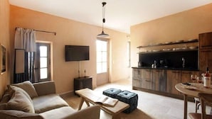 A living room with a sofa, a coffee table, and a TV. There is a kitchen area with cabinets and a dining table with chairs.