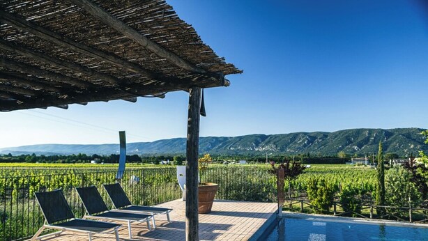 A terrace with a pool, lounge chairs, and a view of a vineyard and mountains.