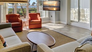 A spacious living room with a large TV, two orange armchairs, a round coffee table, and a view of the ocean through large windows. - 6101 Hampton Place-Modern meets the Beach in this Oceanfront Escape! (Hilton Head Island)