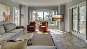 A spacious living room with a gray sofa, two armchairs, a coffee table, and a flat-screen TV mounted on the wall. Large windows provide a view of the outdoors. - 6101 Hampton Place-Modern meets the Beach in this Oceanfront Escape! (Hilton Head Island)