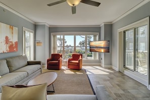 A spacious living room with a gray sofa, two armchairs, a coffee table, and a flat-screen TV mounted on the wall. Large windows provide a view of the outdoors.